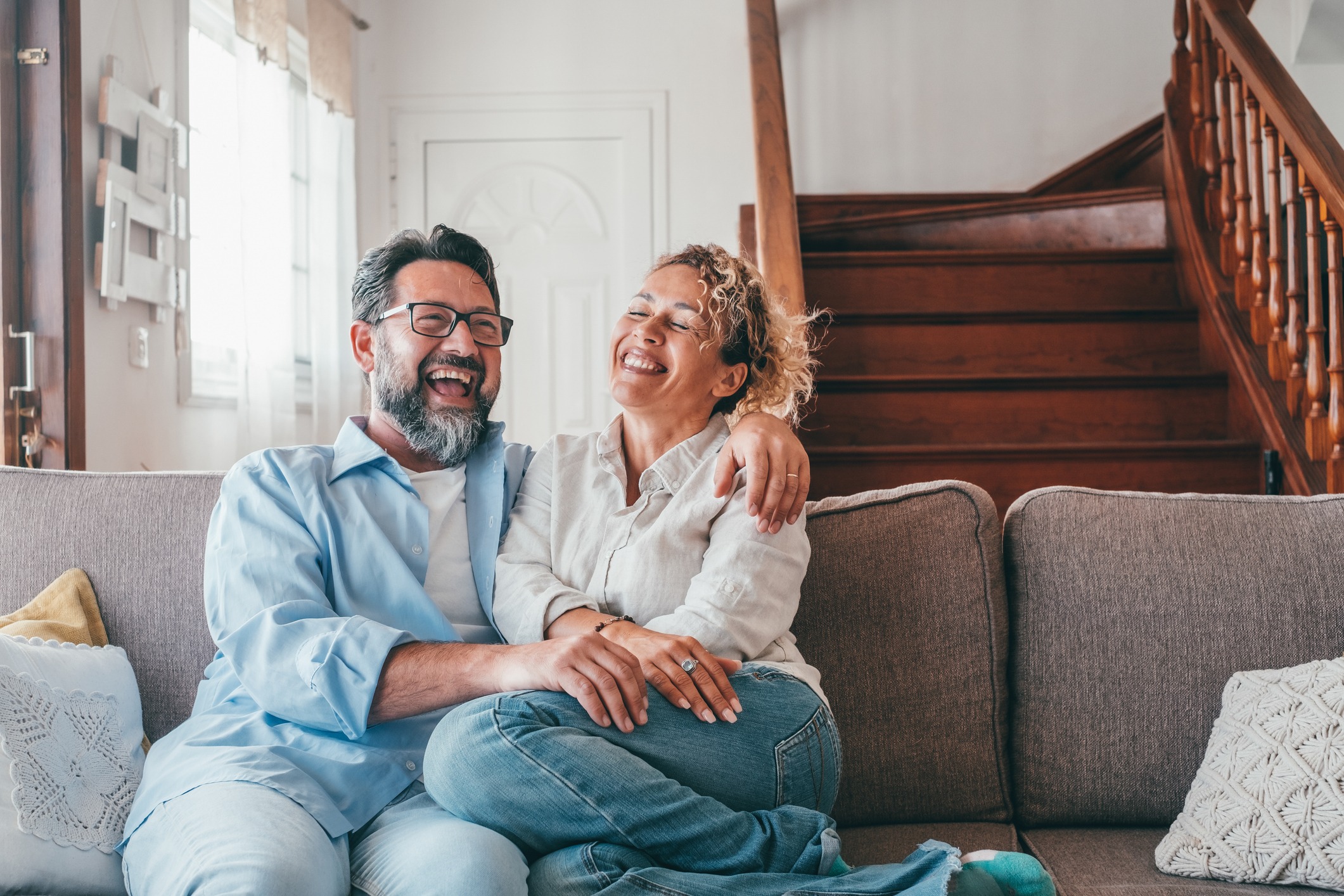 couple laughing while sitting on sofa in the living room. Husband and wife laughing out loud sitting on couch at home. Man and woman spending leisure time at modern apartment